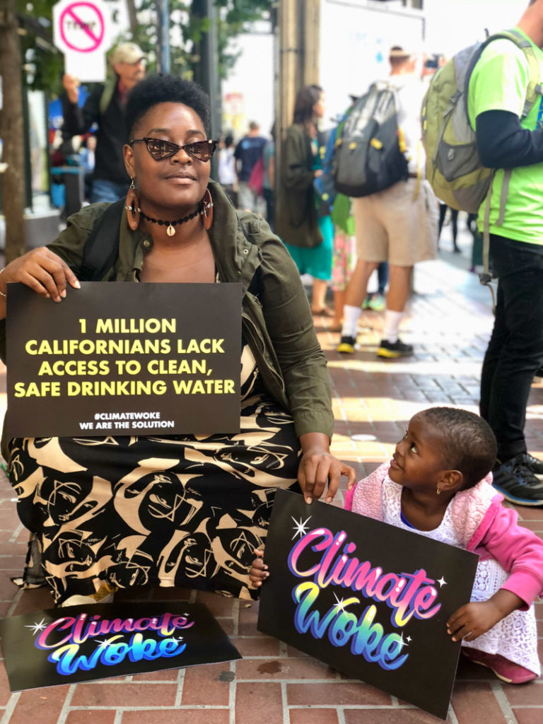 A mother and her small child are holding up Climate Woke signs that say: “1 million Californians lack access to clean, safe drinking water”