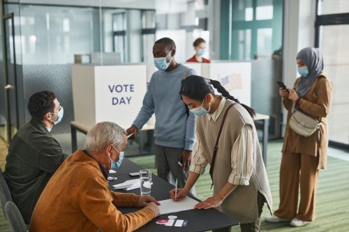 Two people sitting at a registration table, three people waiting to enter voting booths