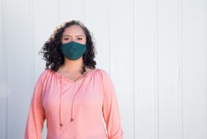 Woman wearing pink shirt and a k95 mask stands in front of a white wall.