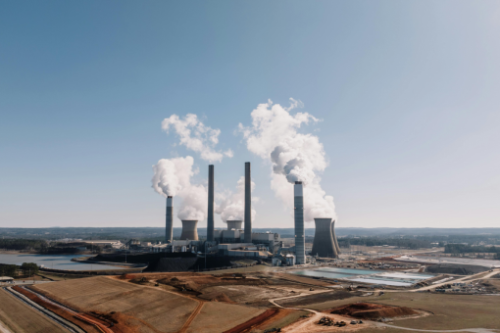 A wide view of a coal-fired power plant with multiple tall smokestacks emitting white smoke into a clear blue sky. The plant is surrounded by open fields, a water reservoir, and industrial structures, emphasizing the industrial and environmental impact of coal energy production.