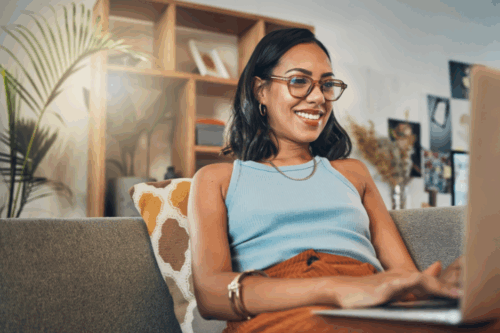 Person of color sits on the couch on her laptop, smiling.