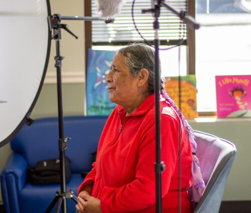 Older woman in a red jacket sits with video equipment in the foreground